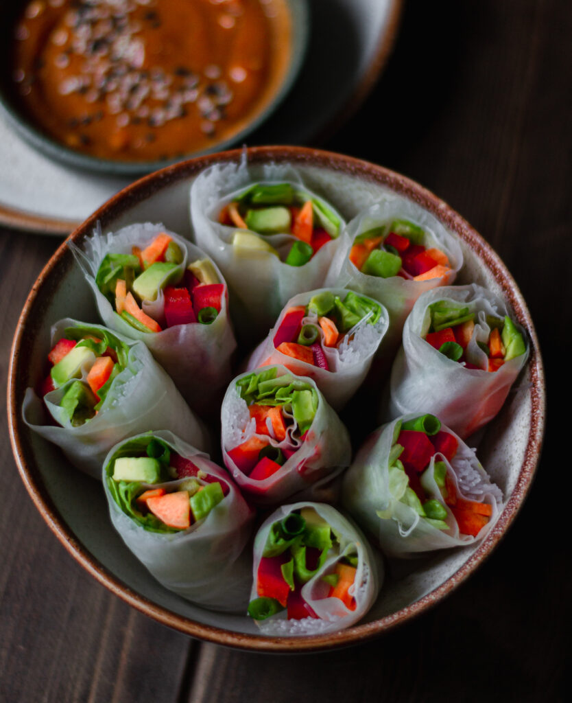 vegetarian spring rolls filled with colourful veggies: avocado, pepper, carrots, spring onion with a side of spicy tahini sauce and sesame seeds
