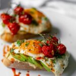 toasted bread with a layer of hummus, avocado slices and fried eggs, topped with roasted cherry tomatoes and sweet chilli sauce